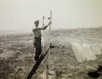 Old black and white photo (1950), of Vernon Zwicker in Country Island. In the background is the windmill used to charge the batteries for the CCG radio.