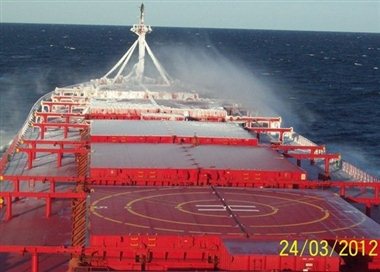 A cargo vessel painted red has white frost on its bow due to ice accumulation on its front structures. The weather seems rather good and the sea calm enough, yet the freezing spray occurs from small wave washes as explained in the paragraphs.